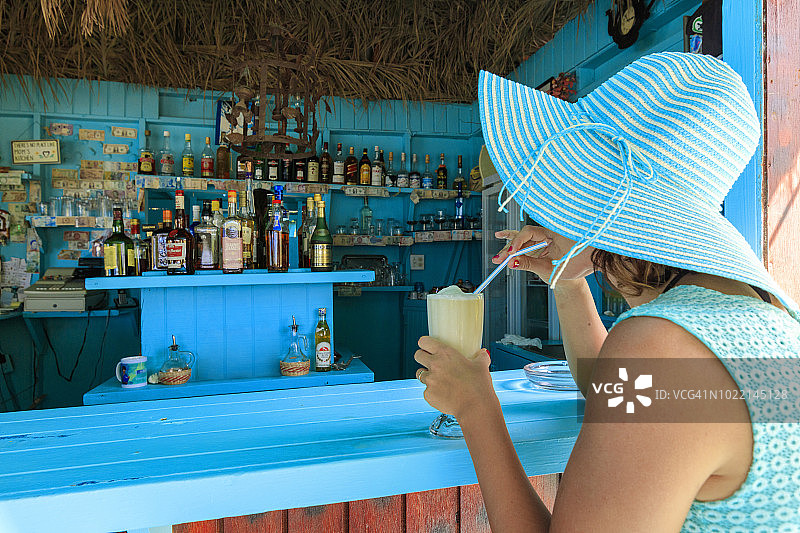 女人戴着帽子和迷人的裙子在安提瓜长湾海滩酒吧喝新鲜水果鸡尾酒图片素材
