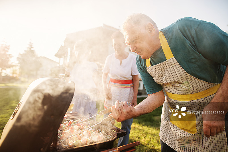 快乐的老人正在检查他的肉在烧烤，而他的家人正在享受在后院图片素材