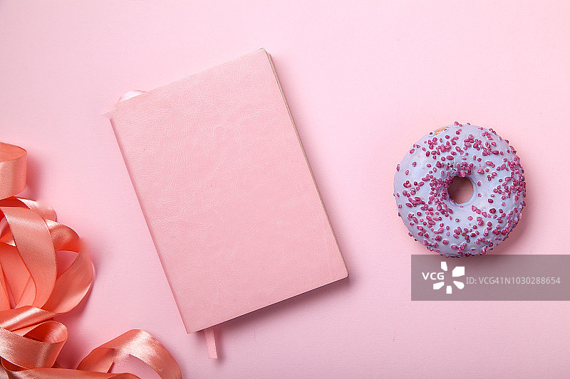 粉红色的笔记本上有闪亮的粉色缎带和粉色背景上的甜甜圈。单色的概念。图片素材