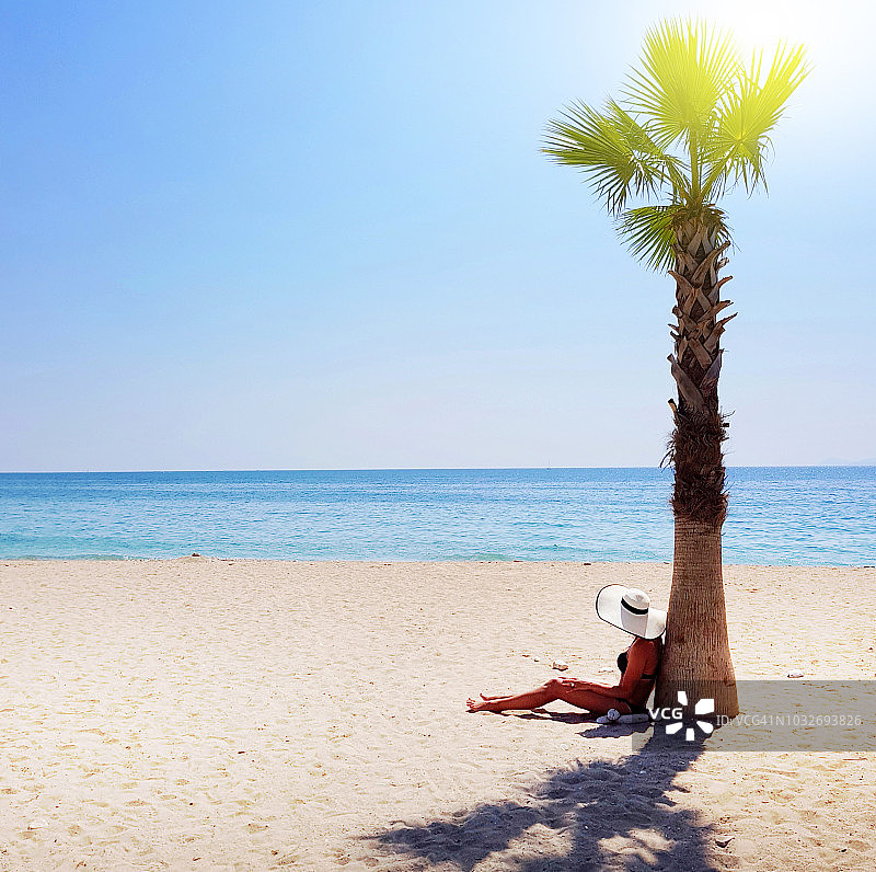 一个戴着白色草帽的女人坐在沙滩上的棕榈树下，看着大海图片素材