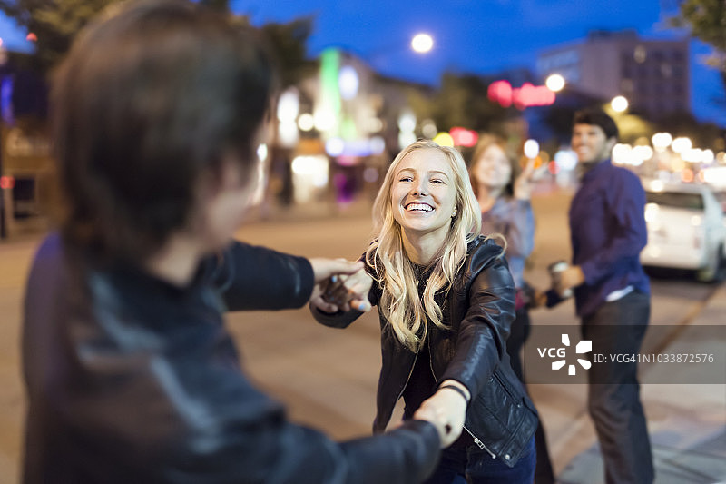 一对穿着黑色皮夹克的年轻夫妇在夜晚的城市人行道上玩耍，背景是朋友们的注视图片素材