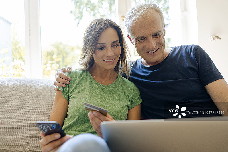 微笑的成熟夫妇坐在家里的沙发上用笔记本电脑和智能手机在网上购物图片素材