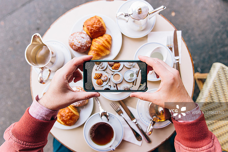 法国巴黎，一名年轻人正用智能手机拍摄路边咖啡馆桌上的牛角面包，享用法式早餐图片素材