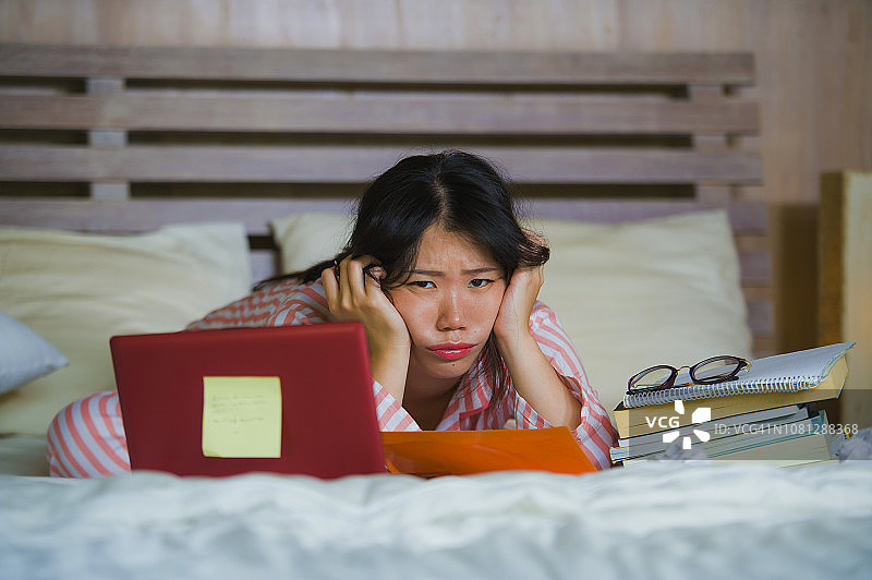 年轻沮丧和疲惫的亚裔美国大学生女孩感到不堪重负和压力准备考试学习与笔记本电脑在压力和在家床上过度工作图片素材