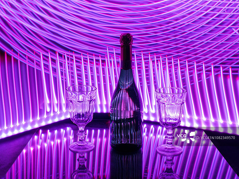 一瓶气泡酒和两个杯子在反光表面。背景中有紫色的线灯。光绘。概念的性质图片素材