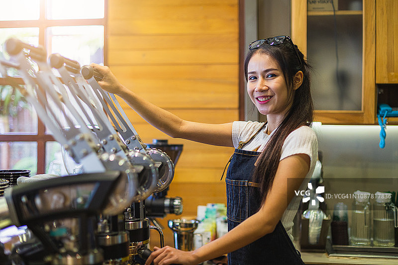 年轻微笑的亚洲女性咖啡师或女性咖啡所有者使用咖啡机，并在咖啡店咖啡厅看着镜头图片素材
