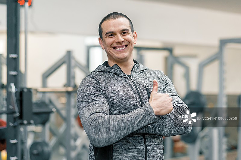 肌肉运动微笑男子的肖像显示在健身房竖起大拇指，英俊的教练看着镜头图片素材