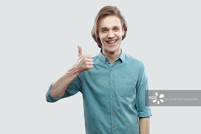 画像满意快乐英俊的长头发金发年轻人在蓝色休闲衬衫站拇指向上和牙齿微笑在相机。图片素材