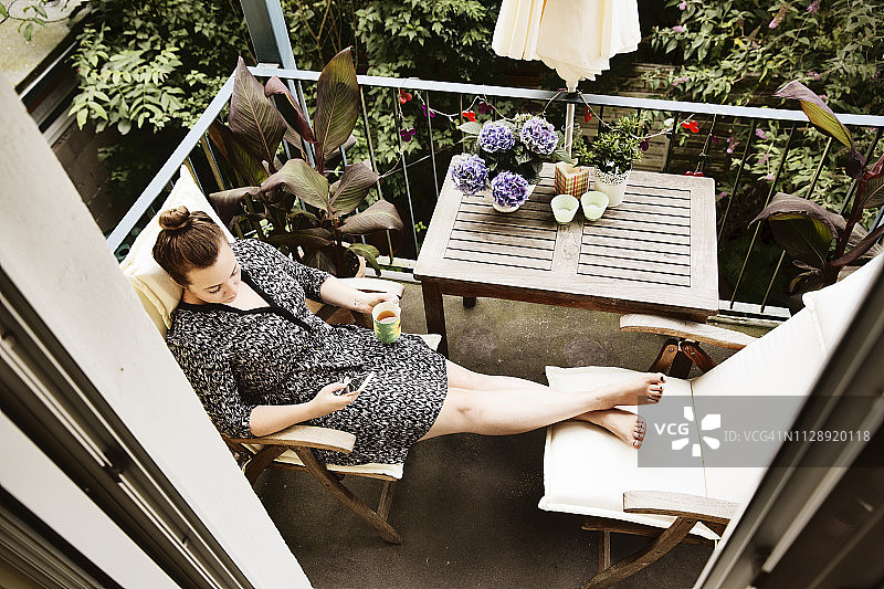 年轻女子坐在阳台上，用智能手机端着一杯茶图片素材
