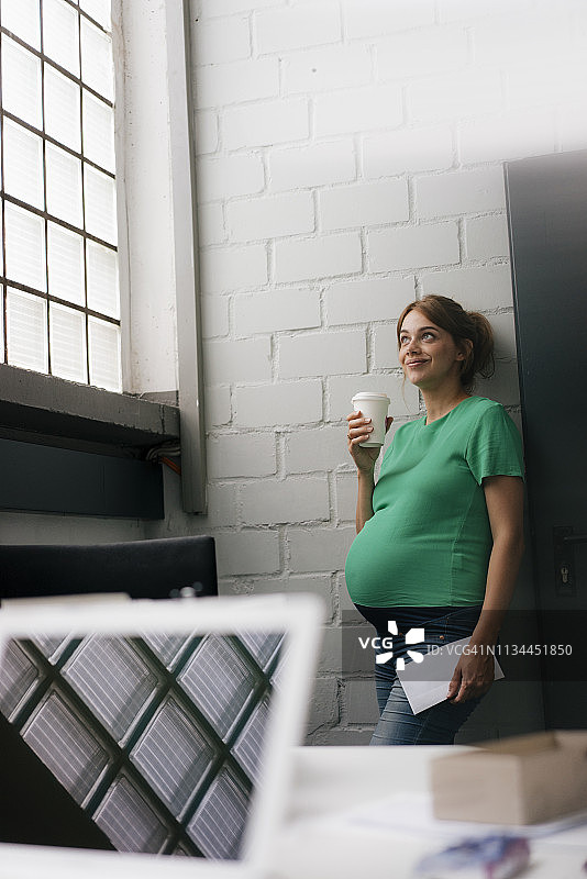 一名孕妇面带微笑，拿着一次性杯子靠在办公室的墙上图片素材