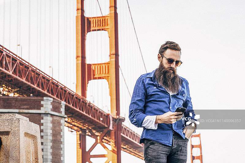 美国加州旧金山，一名留胡子的男子在金门大桥检查智能手机图片素材
