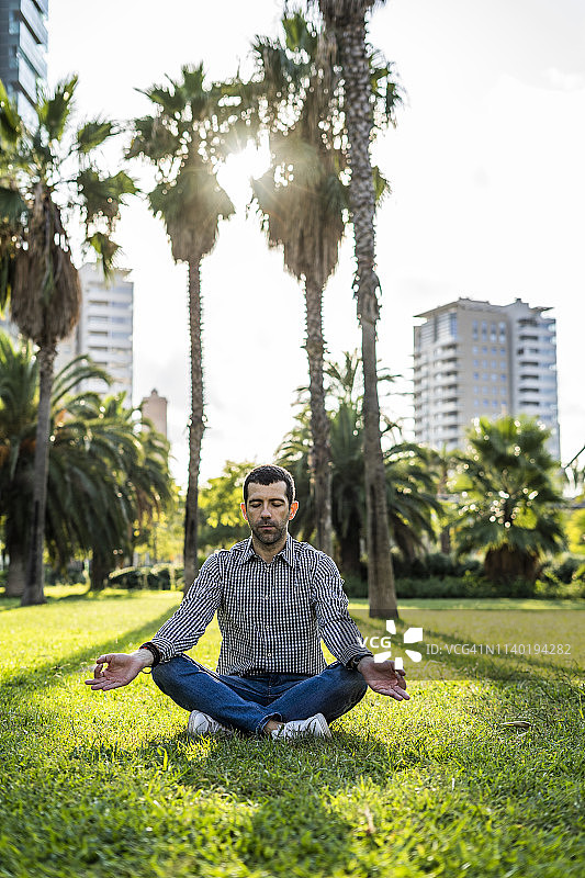一名男子坐在城市公园的草地上做瑜伽运动图片素材