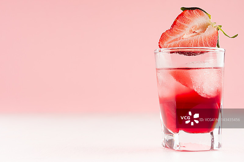 精致的冷红色酒精烈酒，冰块和草莓片在小酒杯近距离白色木桌子和粉粉墙，复制空间。图片素材