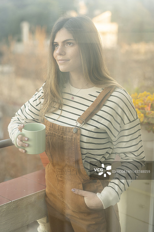 一个微笑的年轻女人的肖像，拿着咖啡杯站在阳台上图片素材