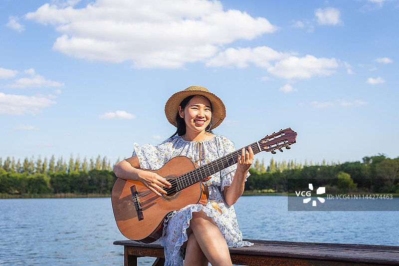 快乐的年轻亚洲女子在湖边和蓝天弹奏古典吉他图片素材