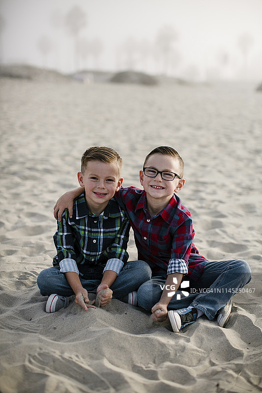 在雾天里，可爱的兄弟坐在沙滩上的微笑肖像图片素材