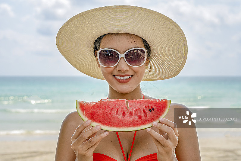亚洲女孩短发和性感的身体谁穿着蓝色比基尼和漂亮的帽子。暑假在美丽的海滩上，她抱着西瓜，开心地吃着西瓜图片素材
