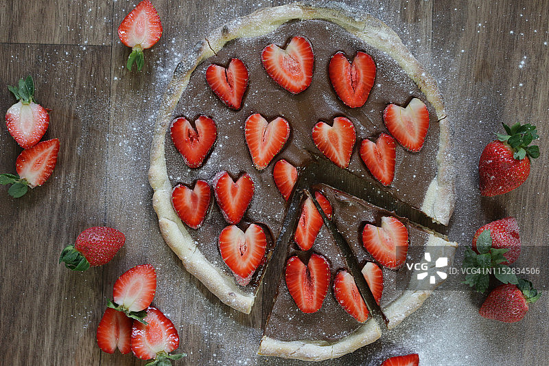 这是一个用草莓和糖霜自制的巧克力披萨，烤披萨的外皮上涂满榛子巧克力，上面是浪漫的红色心形草莓/草莓水果，可以切成薄片当布丁吃图片素材