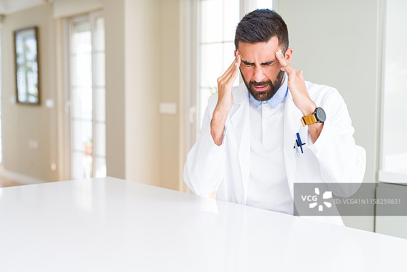 英俊的西班牙医生或治疗师穿着医疗外套在诊所的手在头上疼痛，因为压力。遭受偏头痛。图片素材