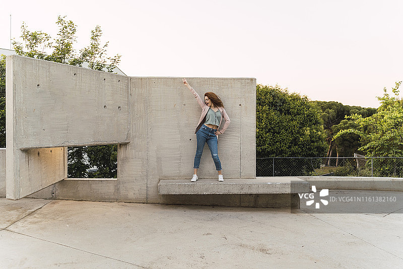 年轻的红发女人，抬起手臂站在水泥长凳上图片素材