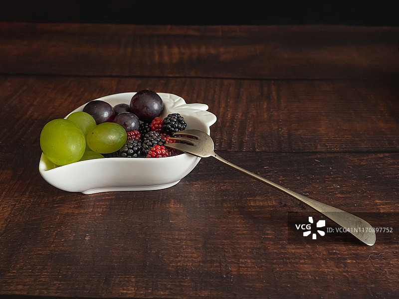 秋天的水果:白葡萄、黑葡萄和黑莓放在一个心形的瓷碗里，旁边放着一把旧羊驼叉图片素材
