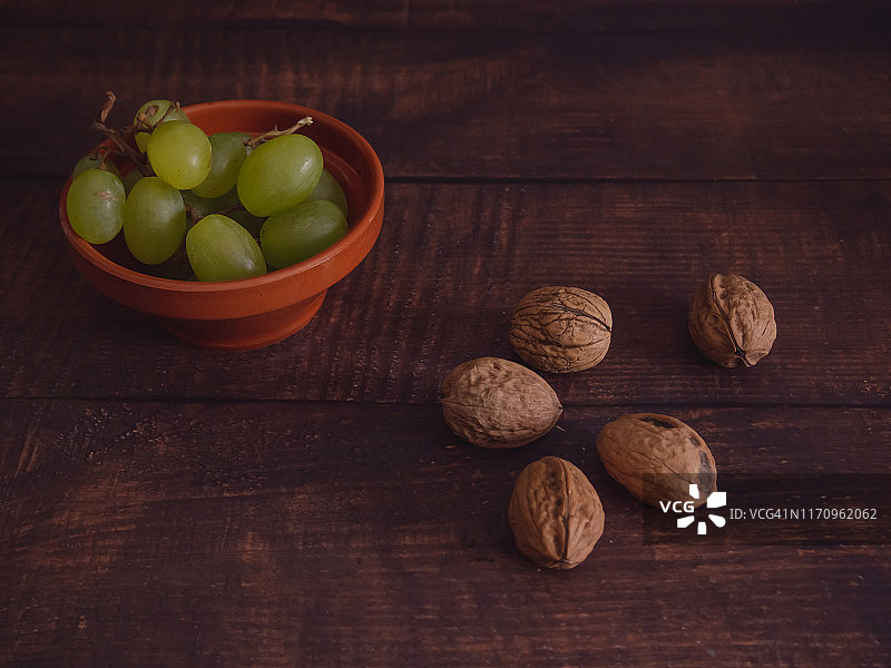 秋天的水果:白葡萄放在一个陶碗里，坚果放在一张旧的深色木桌上图片素材