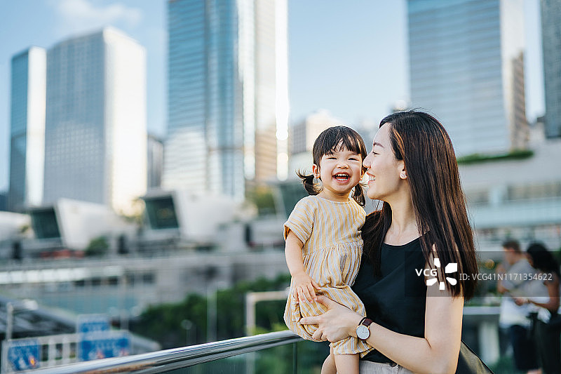 年轻的亚洲母亲和可爱的小女儿在市中心的城市露台上快乐地微笑，对着现代摩天大楼图片素材