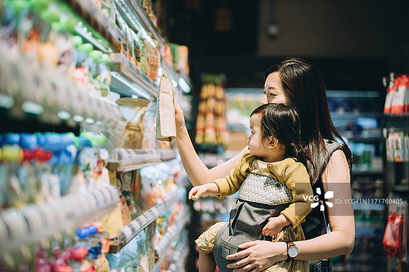 年轻的亚洲母亲带着小女儿在超市购物，选择有机婴儿食品图片素材