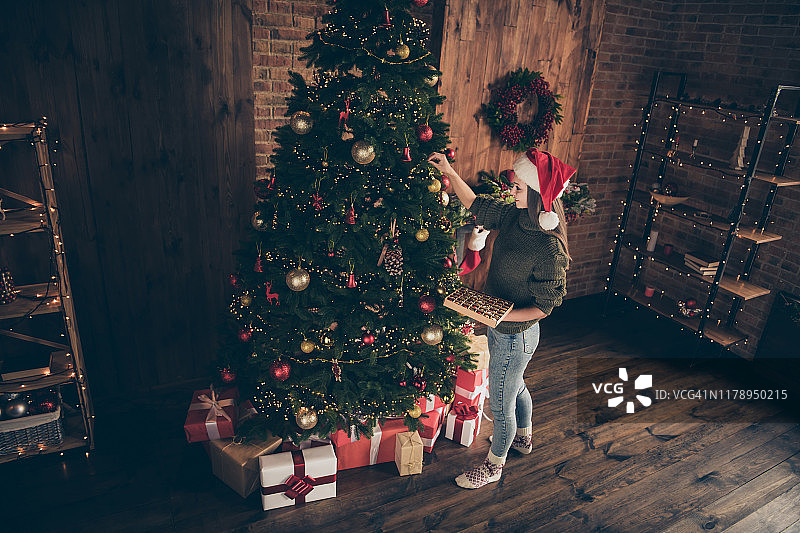 上面的高角度照片正面的黑发女孩手在常青树上的小玩意准备圣诞庆祝派对戴圣诞老人的帽子在房子里与新年装饰灯室内图片素材