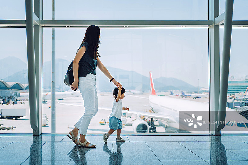 快乐的年轻亚洲母亲牵着可爱的小女儿通过窗口看飞机在机场等待离开图片素材