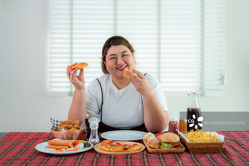 亚洲超重女性拿着披萨吃着桌子上的许多垃圾食品，不健康的营养和暴饮暴食。她吃垃圾食品看起来很开心。饮食不健康的生活方式。图片素材