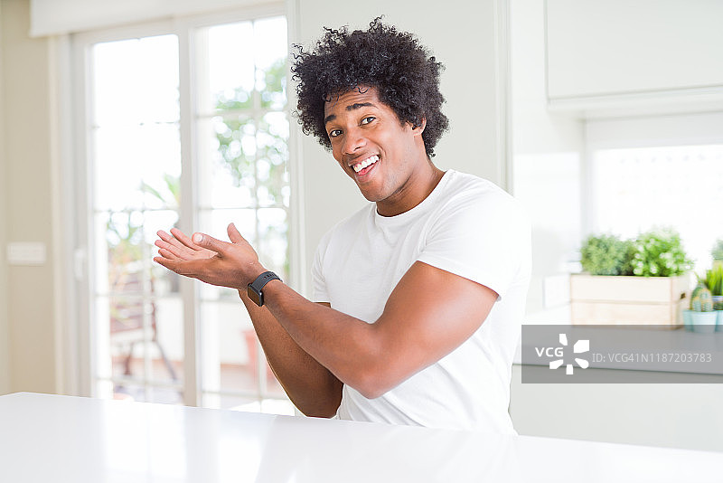年轻的非洲裔美国人穿着休闲的白色t恤坐在家里指着一边，双手张开的手掌显示拷贝空间，呈现广告微笑兴奋快乐图片素材