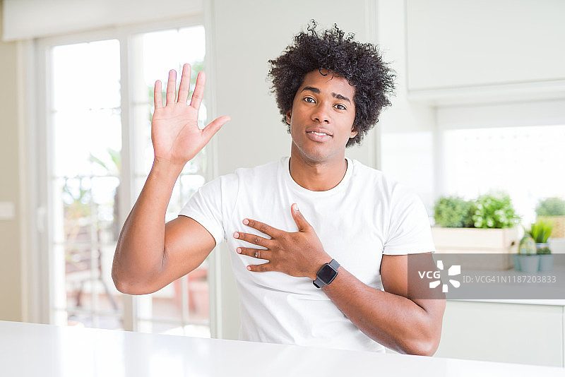 一名年轻的非洲裔美国男子，穿着休闲的白色t恤坐在家里，手放在胸前和摊开的手掌上宣誓，作出忠诚承诺的誓言图片素材