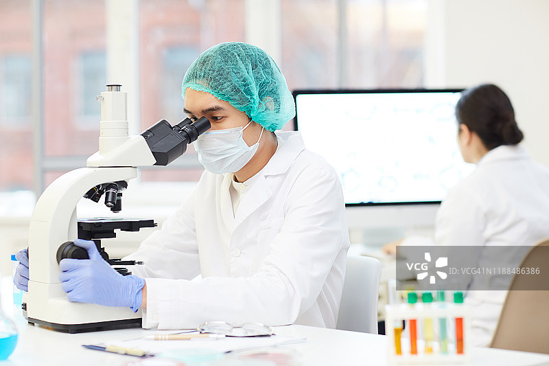 严肃专注的年轻亚洲男性科学家戴着无菌帽和口罩坐在桌子旁，通过显微镜分析实验样品图片素材