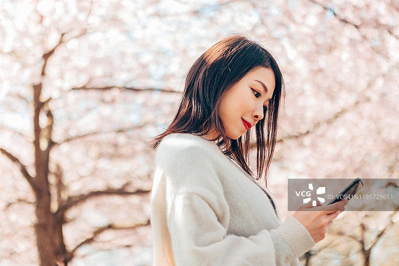 美丽和愉快的年轻女子在花树下使用手机的肖像图片素材