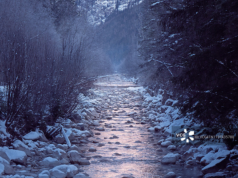 一条雾蒙蒙的山川流过一片白雪皑皑的景色。树木和岩石被雪覆盖，创造了一个完美的冬季宁静的景象。图片素材