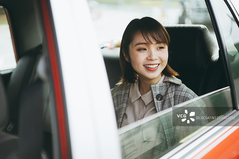 年轻的亚洲女子坐在出租车后座，看着窗外，微笑着在城市通勤图片素材