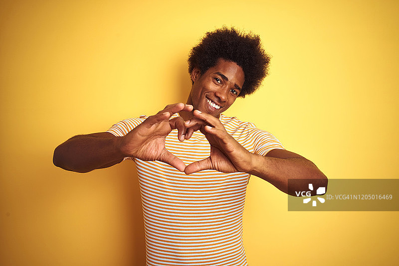 美国男子与非洲式头发穿条纹t恤站在孤立的黄色背景微笑在爱做心形符号形状的手。浪漫的概念。图片素材