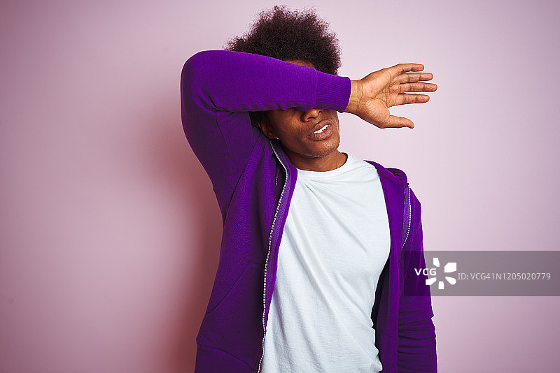 年轻的非洲裔美国人穿着紫色的运动衫站在孤立的粉红色背景覆盖手臂，看起来严肃和悲伤的眼睛。失明，隐藏和排斥的概念图片素材