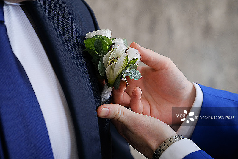 新郎的朋友正在整理扣孔上的钮扣。一个穿着白色衬衫和优雅的蓝色夹克的年轻人，在他的背心口袋里有一朵花。婚礼的主题。图片素材