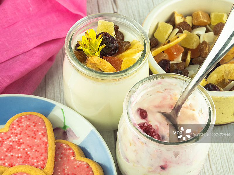 上图是一杯浆果酸奶，一杯水果干酸奶，一个水果干碗和一个心形饼干盘子图片素材