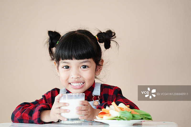 亚洲可爱的小女孩在早上喝着健康的新鲜牛奶和新鲜的绿色蔬菜沙拉。牛奶含有蛋白质、维生素和钙，对儿童的健康非常有益。图片素材