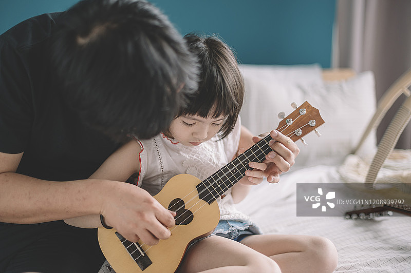 周末，在卧室的床上，一个亚洲华人的小女儿在向她的父亲学习弹奏尤克里里琴图片素材