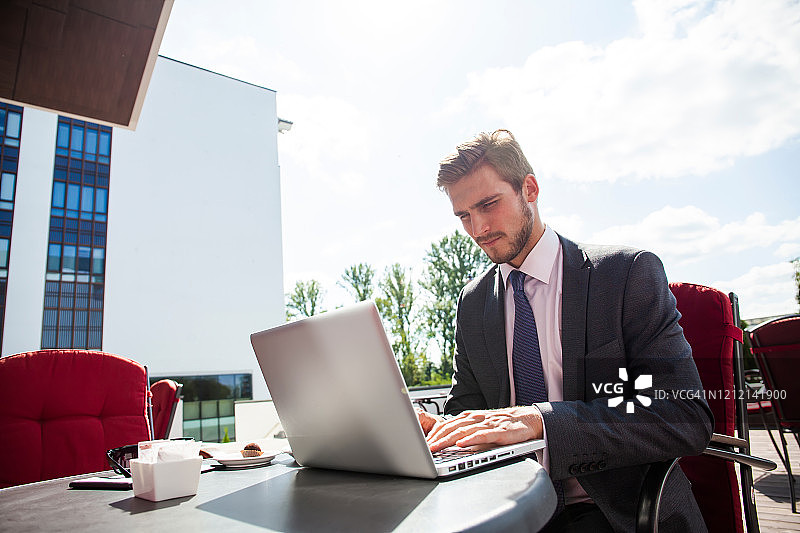 英俊的商人穿着西装，使用现代笔记本电脑在户外，成功的经理工作在咖啡馆休息和搜索信息在他的笔记本电脑上。图片素材