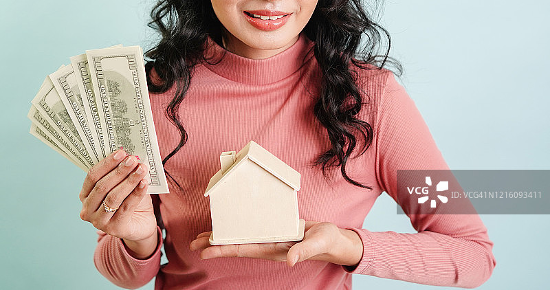美丽的年轻女子持有美元钞票和房子模型在蓝色的背景-房地产贷款融资和购买房子的概念。图片素材