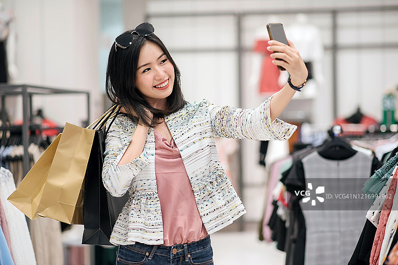 亚洲华人女性手握手机，在服装店微笑自拍的肖像图片素材