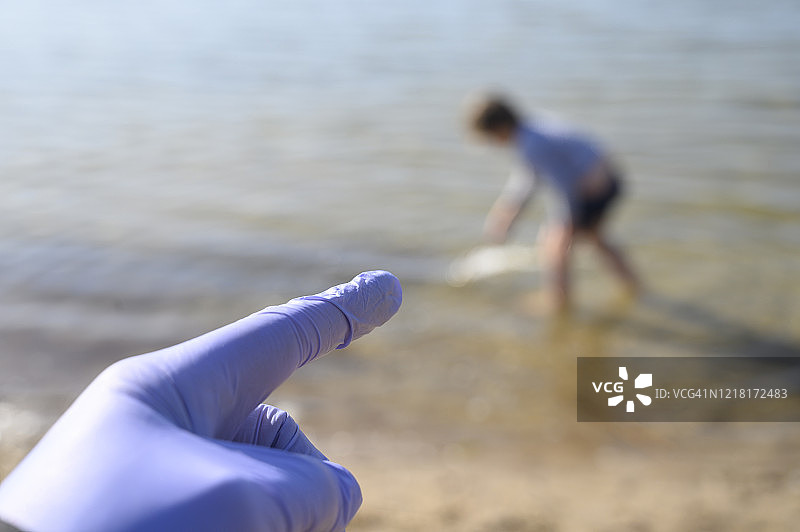 手戴保护手套，食指指向一个小男孩(认不出来)在湖边的沙滩上玩耍。德国。图片素材
