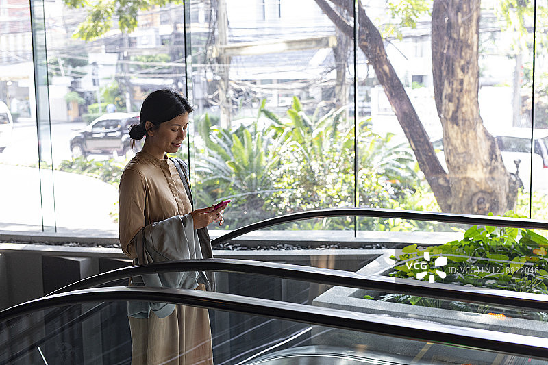 保持联系:快乐的亚洲女人在乘坐自动扶梯时用手机发短信图片素材