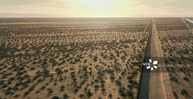 一个阳光明媚的下午，在加州/墨西哥沙漠中，远处的群山为背景，一条与墨西哥和美国之间的钢条边境墙平行的土路图片素材