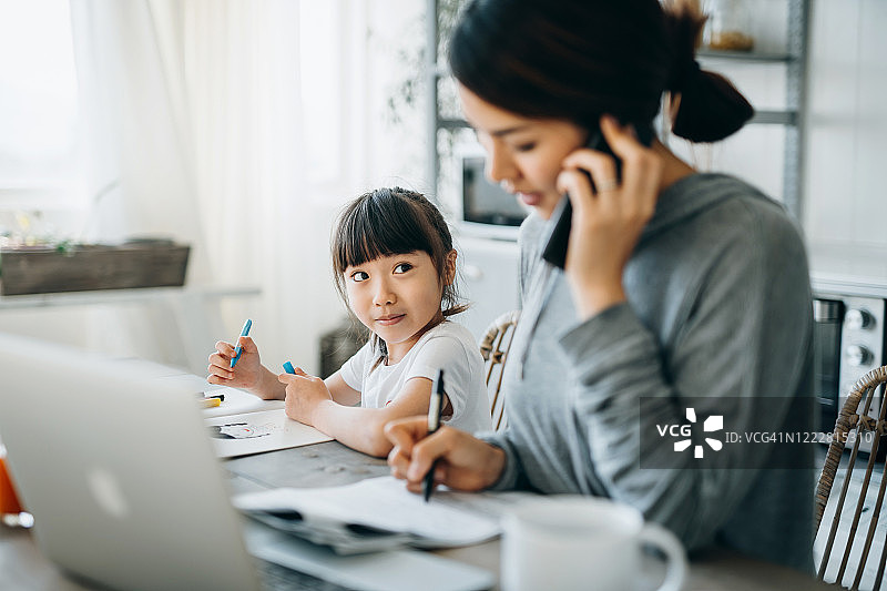 年轻的亚洲母亲在家通过笔记本电脑和电话工作，而小女儿在家里学习。她用平板电脑上在线课程，在家做作业图片素材